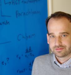 A portrait of Greg Fournier in a grey sweater standing next to a blue whiteboard with black writing.